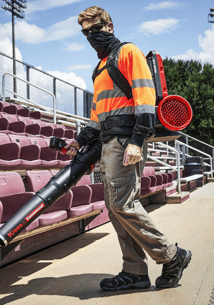 A worker in high-visibility safety gear and a face mask operates a Kress Commercial backpack leaf blower to clear debris from stadium bleachers